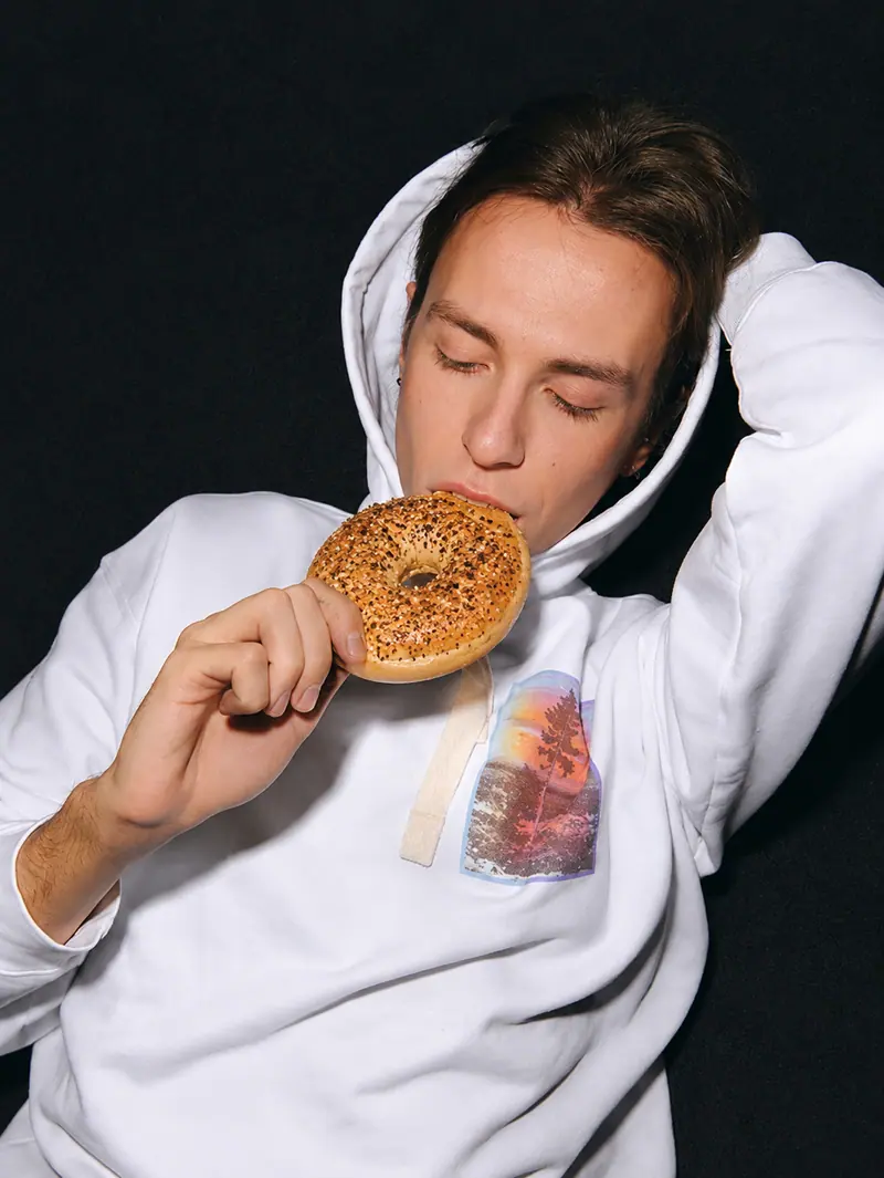 Person in a white hoodie biting a bagel